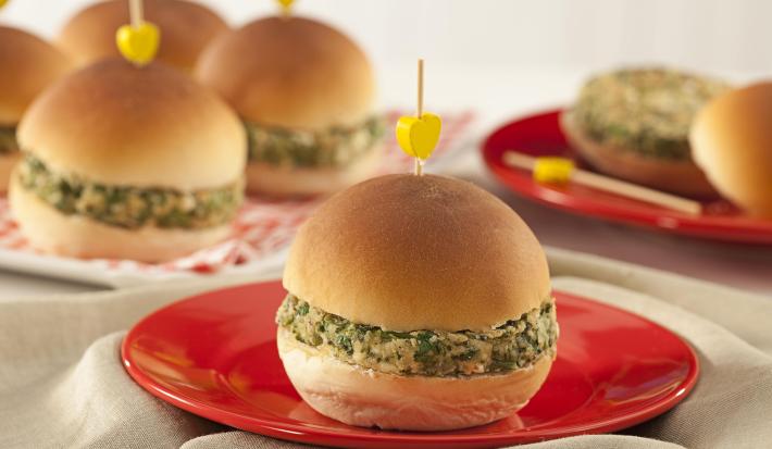 A foto contém diversos pães com hamburguês e palitos com uma bolinha amarela por cima. Um prato vermelho com um pão com hambúrguer à frente