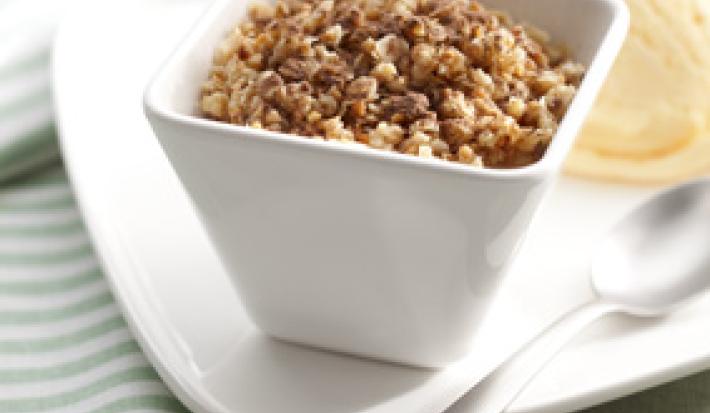 Fotografia em tons de verde em uma bancada de madeira verde clara, um pano listrado com verde, um prato branco, um potinho branco quadrado com o crumble de maçã dentro dele. Ao lado, uma bola de sorvete de creme.