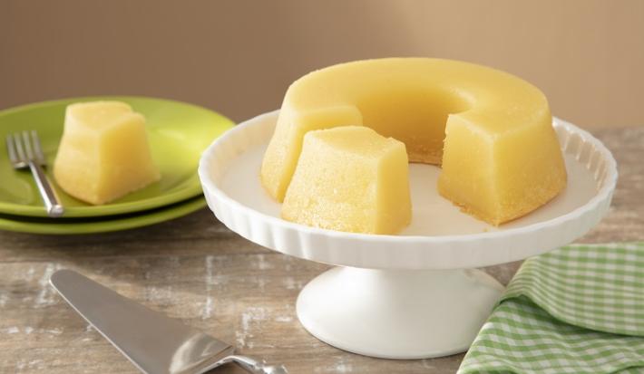Foto em tons de amarelo da receita de bolo souza leão de pernambuco servida em uma base de porcelana alta circular com um prato de porcelana verde com um pedaço da receita. Ao lado há um paninho verde quadriculado e uma espátula prateada
