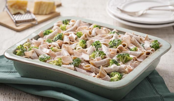 Fotografia em tons de verde em uma bancada de madeira com um refratário retangular de cerâmica com o macarrão tipo penne com brócolis e molho de creme de leite dentro dele.