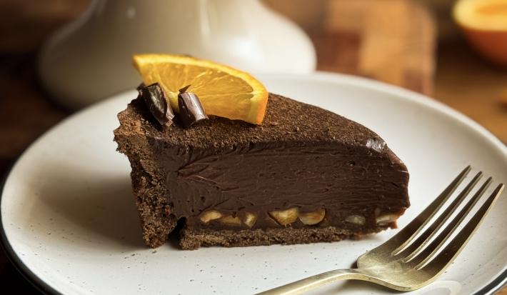 Fotografia de uma fatia de torta de chocolate e laranja com avelã em um prato pequeno raso branco. Ao lado da fatia tem um garfo apoiado no prato também.