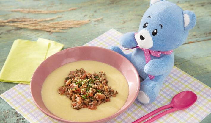 Foto da receita de Polenta Mole com Molho de Carne Moída. Observa-se um potinho rosa bebê com a polenta e a carne dentro, sobre um paninho quadriculado colorido. O ursinho Bo está do lado direito.