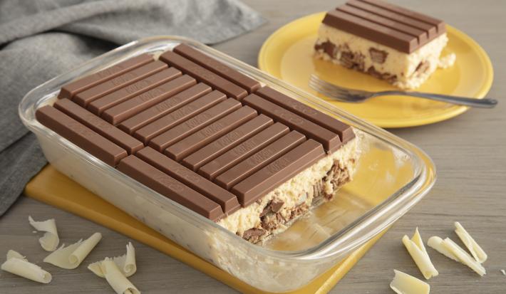 Fotografia em tons de amarelo em uma bancada de madeira com um paninho cinza. Ao centro, uma tábua amarela com um refratário de vidro e a lasanha de chocolate kitkat dentro cortada. Ao lado, um prato amarelo com um pedaço da lasanha de chocolate.