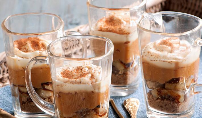 4 tacinhas transparentes com o doce banana com brigadeiro de caramelo e polvilhadas com canela. As 4 estão dentro de um cesto marrom com um pano azul.