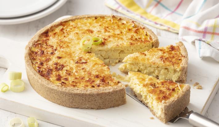 Fotografia em tons de branco de uma bancada branca com uma tábua branca, sobre ela o quiche com uma espátula tirando uma fatia. Ao fundo dois pratos brancos e um paninho branco.