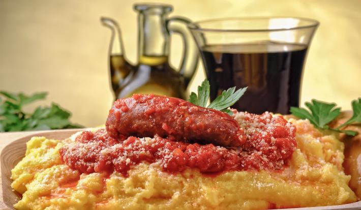 Fotografia de uma polenta com molho de tomate, cebolas e linguiça, que está dentro de um prato fundo de madeira. Por cima, queijo ralado e algumas folhas de salsinha. No fundo, muito azeite em um recipiente de vidro na frente de uma parede amarela.