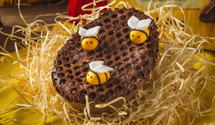 Foto aproximada de um ovo de chocolate de colher decorado com modelo de favo de mel e abelhas de mentira, sobre uma bancada com palha, tecido amarelo e uma barra de chocolate Garoto ao fundo
