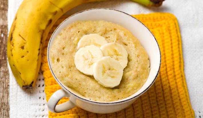 Fotografia em tons de amarelo com uma xícara de café ao centro. Dentro da xícara existe uma receita de bolo de banana. Ao lado existe uma banana inteira em cima de um pano amarelo