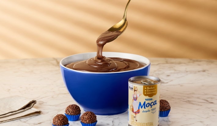 Fotografia mostra um pote cheio de brigadeiro. Alguns brigadeiros em forminhas azuis em volta e a lata de leite moça.
