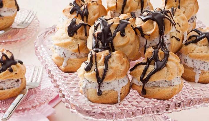 Fotografia em tons de rosa de uma bancada de rosa com um suporte de vidro com profiteroles. Ao lado dois pratos de vidro pequenos com um profiteroles e um garfo.