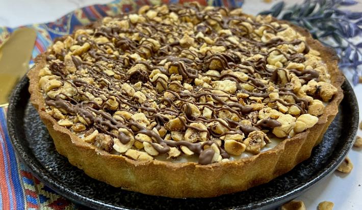 Foto da receita de Torta Nocciolíssima. Observa-se uma torta inteira decorada com avelãs e creme de chocolate sobre um prato de cerâmica redondo preto.