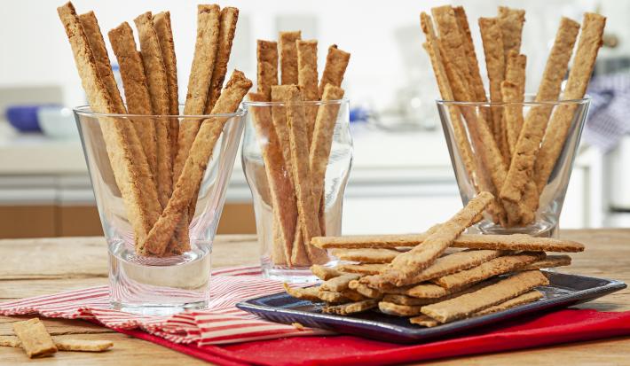 Fotografia em tons de pardo e vermelho de uma bancada de madeira marrom vista de frente, contém 3 copos transparentes e cada um contém biscoitos de cebolas em tiras. Ao lado uma travessa preta com mais biscoitos em tiras e abaixo um pano vermelho.