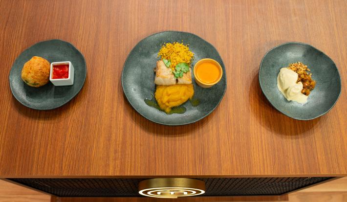 Fotografia em tons de marrom com três pratos cinzas ao centro. Dentro de um prato existe uma sobremesa de sorvete com amendoim, de outro existe um peixe e em outro uma entrada frita acompanhada de um molho de cor vermelha.