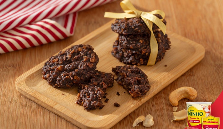 Cookies de cacau com castanhas sobre tábua de madeira, alguns quebrados, com castanhas ao redor e pano listrado ao fundo.