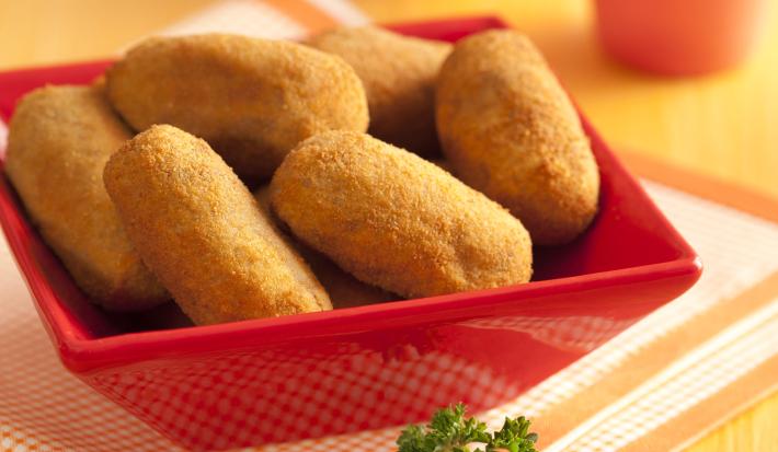 Fotografia em tons de amarelo e vermelho de uma bancada de madeira com um pano dobrado quadriculado branco e laranja, um recipiente vermelho quadrado com vários bolinhos fritos e ao fundo dois copos rosa.