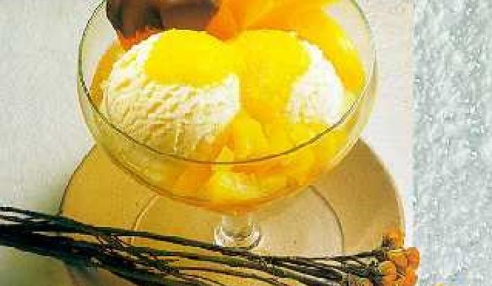 Fotografia em tons de amarelo em uma bancada de madeira, um pano bege, um prato redondo raso bege com uma taça de vidro com o sorvete de creme e pêssegos em calda dentro dela. Ao lado, flores amarelas e laranjas.