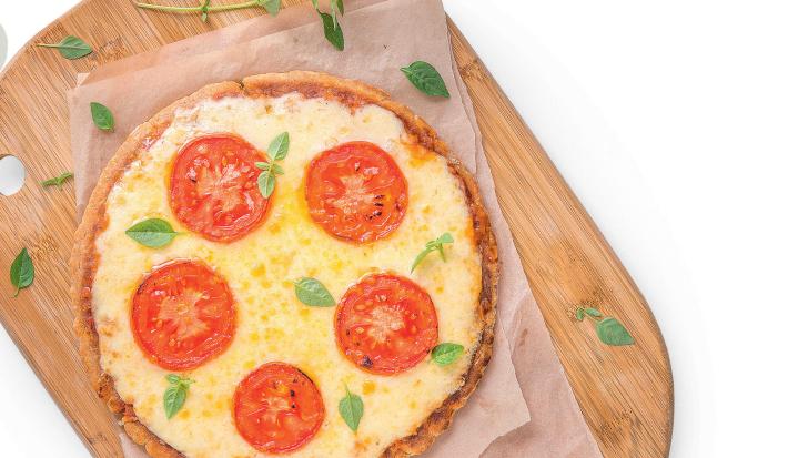 Fotografia em tons de amarelo e vermelho em uma mesa branca com uma tábua de madeira, um pano bege e uma pizza média de queijo com tomate em cima dela.