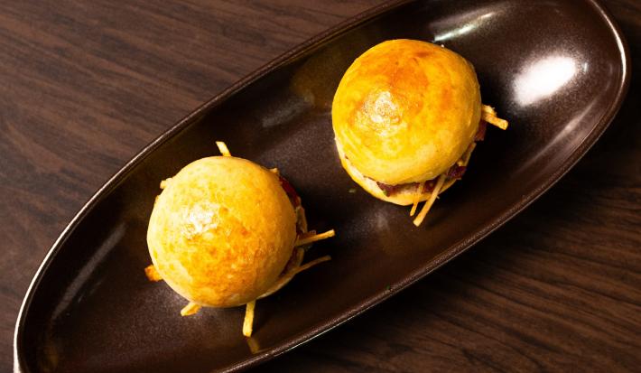 Fotografia em tons de marrom com um prato marrom escuro ao centro. Dentro do prato existe dois pães recheados com um tartare de carne, picles e batata palha