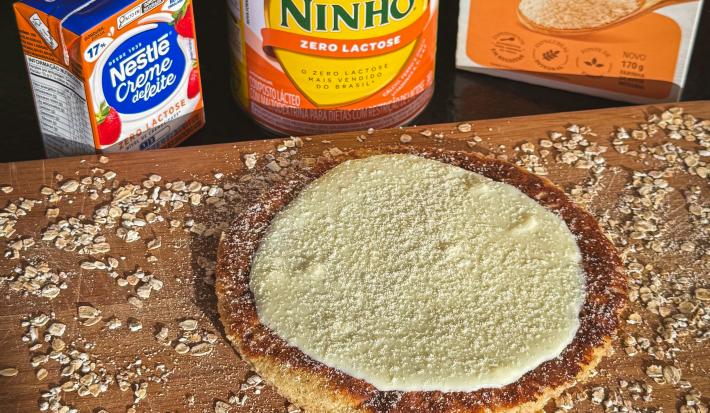 Fotografia em tons claros de uma tábua de madeira salpicada com farelo de aveia e tem uma panqueca de aveia com creme de ninho por cima e ninho salpicado. Atrás da tábua tem aveia e produtos zero lactose nestlé.