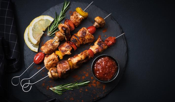 Fotografia vista de cima de três espetinhos, um do lado do outro, de carnes intercaladas com legumes. Os espetos estão sobre um apoio preto redondo, ao lado de duas fatias de limão siciliano, um molho vermelho em um recipiente menor preto e alecrim.