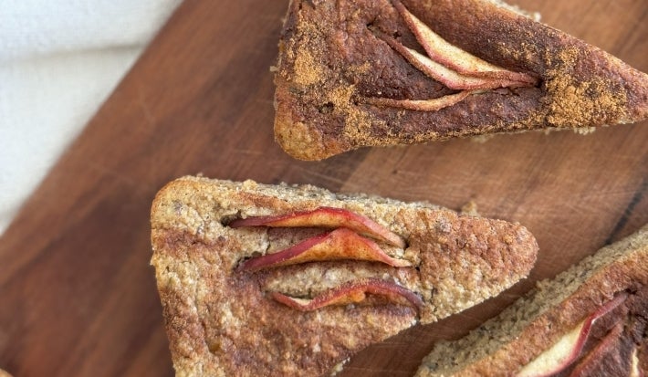Fotografia em tons de marrom com uma tábua de madeira ao centro. Em cima da tábua existe fatias de um bolo de maçã finalizado com canela.