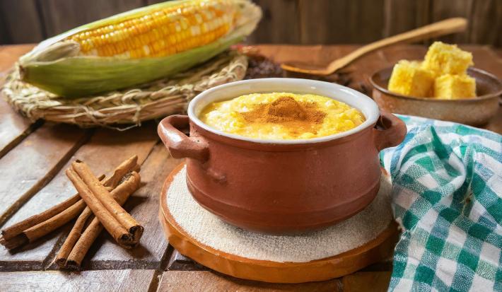 Fotografia de uma mini panela de barro apoiado em um apoio branco sobre uma mesa de madeira. Dentro da panelinha tem uma receita de canjica de milho verde com canela por cima. Ao redor tem paus de canela e um milho verde inteiro.