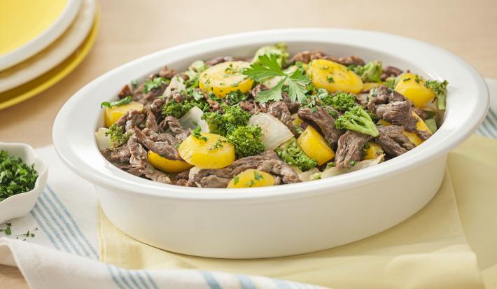 Fotografia em tons de amarelo em uma bancada de madeira clara com um pano amarelo e outro branco com listras azuis e um recipiente oval fundo com as tirinhas de carne com legumes dentro dele. Ao fundo, pratos rasos amarelos e um potinho com salsinha.