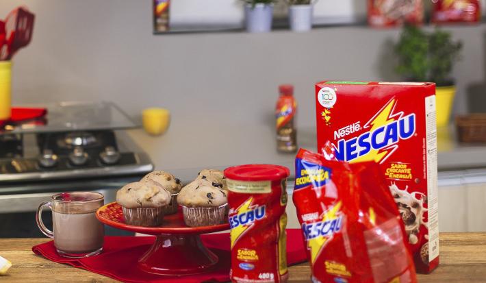 Foto vista de frente para um bancada com produtos Nescau à direita, mais ao centro um prato vermelho alta com quatro Muffins Crocantes de Nescau e à esquerda uma caneca com leite e Nescau.