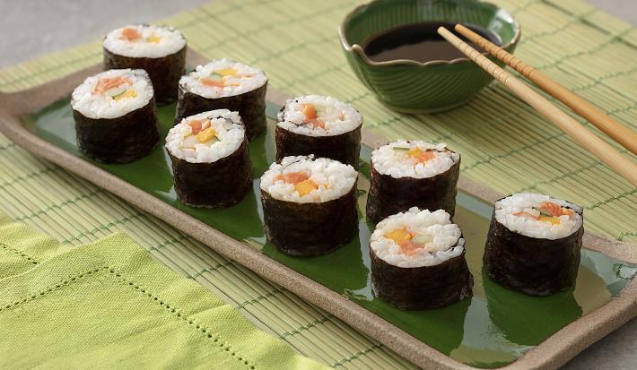Foto em tons de verde da receita de hossomaki básico servida em 9 porções dispostas sobre uma régua de porcelana verde escura com borda marrom. Ao lado há dois hashis sobre um recipiente verde escuro com shoyo