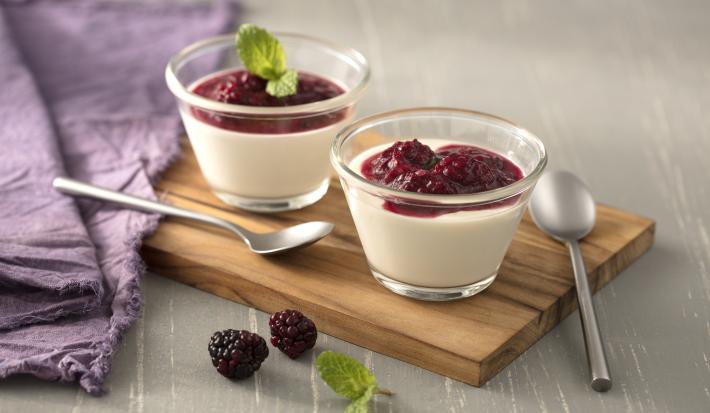 Fotografia em tons de branco e lilás, ao centro dois potinhos com creme branco e molho de framboesa e duas colheres sobre tábua de cozinha, ao lado guardanapo lilás, tudo sobre bancada em tom de lilás.