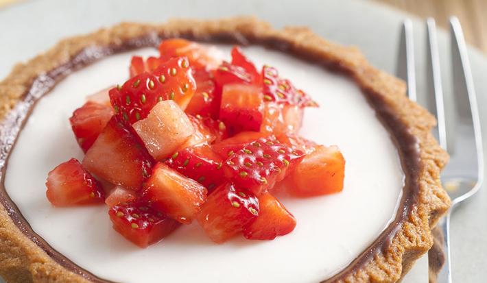 fotografia em tons de branco e vermelho tirada de um prato redondo branco que contém uma mini-torta com creme branco e pedaços de morangos com um garfo ao lado para servir.