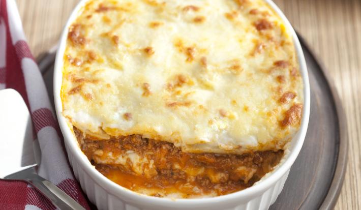 Fotografia em tons de branco e marrom, ao centro travessa branca com lasanha gratinada sobre tábua de cozinha, lado esquerdo guardanapo xadrez branco e vermelho, em bancada de madeira. Ao fundo, pedaço de lasanha em prato vermelho.
