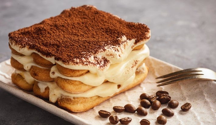 Fotografia em tons claros de uma mini tiramisú clássica sobre um apoio retangular claro, ao lado tem alguns grãos de café inteiros e um garfo apoiado. O recipiente está sobre uma mesa cinza.