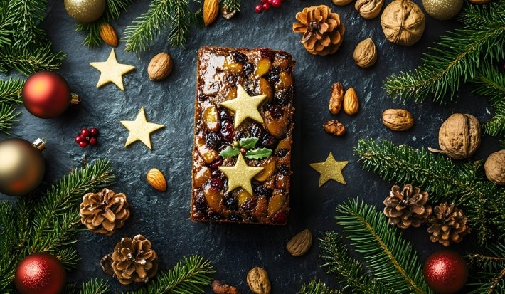 Fotografia de um bolo inglês decorado para as festas, com frutas secas, nozes e estrelas douradas, cercado por ramos de pinheiro e enfeites natalinos.
