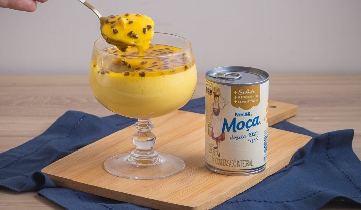 Foto da receita de Mousse de maracujá feita com o suco concentrado da fruta, servida em taças de vidro, com uma decoração de calda de maracujá com sementes, sobre uma tábua de madeira e ao lado de uma lata de Leite Moça