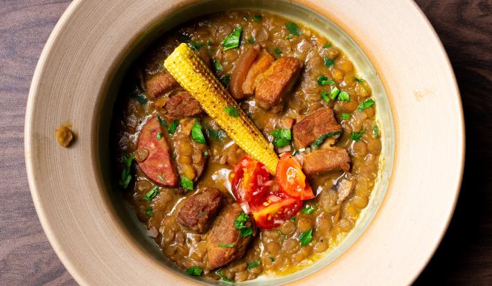 Fotografia em tons de marrom com um prato cinza ao centro. Em cima do prato existe uma receita de lentilha com carne de porco, tomates e mini milhos