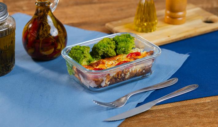 A fotografia mostra uma marmita de vidro com pedaço de torta de frango cremosa, com crosta dourada, tomates-cereja e muçarela derretida. Ao lado, brócolis, em um tom vibrante de verde, compondo um prato equilibrado e saboroso.