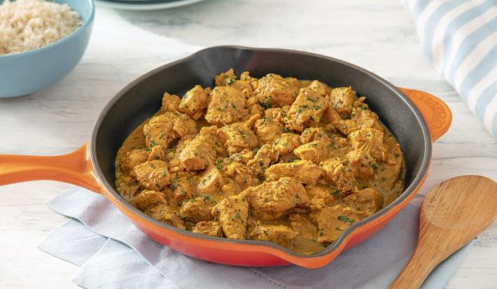Foto da receita de frango ao curry sem lactose servida em uma frigideira grande laranja sobre uma mesa clara com um pano azul embaixo e uma colher de madeira ao lado