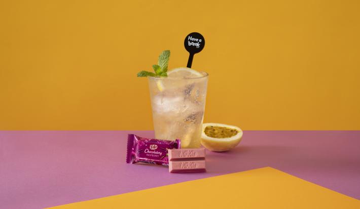 Foto da receita de Soda Italiana Passion Fruit and Lemon. Observa-se um fundo amarelo e roxo com um copo alto no centro, cheio de gelo, com uma rodela de limão siciliano. Meio maracujá decora a foto à direita, assim como folhas de hortelã no copo.