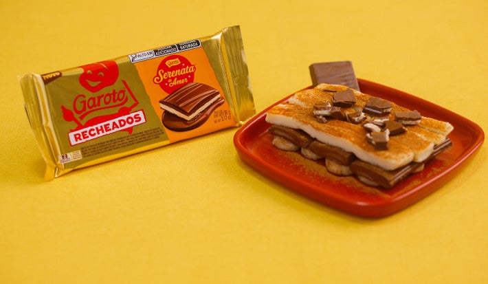Sobremesa com banana, creme e chocolate recheado em prato vermelho, com embalagem Garoto ao lado.
