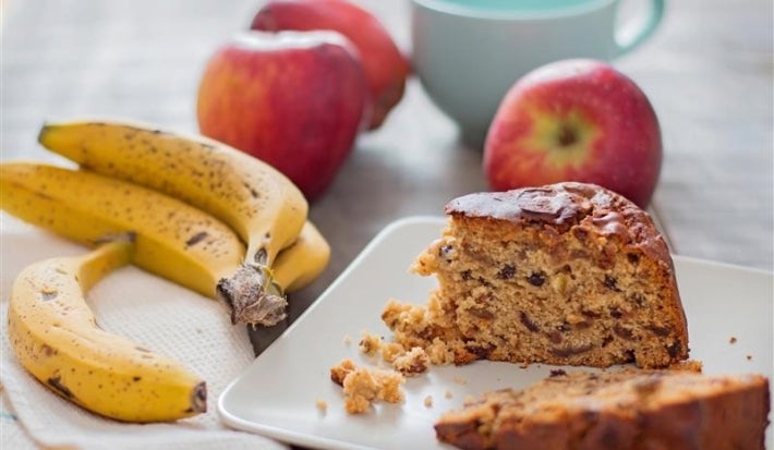 Fotografia de uma fatia de bolo de frutas em um prato branco, acompanhada de bananas e maçãs. O cenário é casual e acolhedor, com uma xícara azul ao fundo, sugerindo um lanche caseiro.