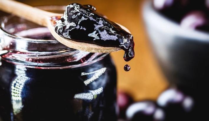 Fotografia de uma colher de madeira sobre um recipiente de vidro com geleia de jabuticaba. A colher também carrega um pouco da geleia, que cai aos poucos na ponta.