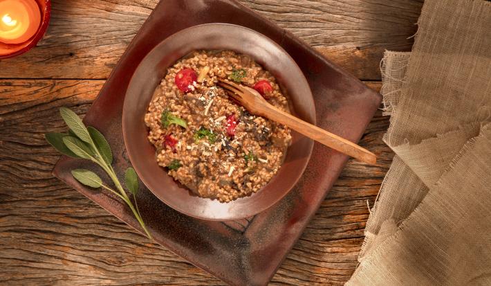 Foto em tons de marrom da receita de risoto ao funghi com aveia corte rústico servida em um prato fundo marrom sobre uma base retangular de mesma cor em cima de uma mesa de madeira