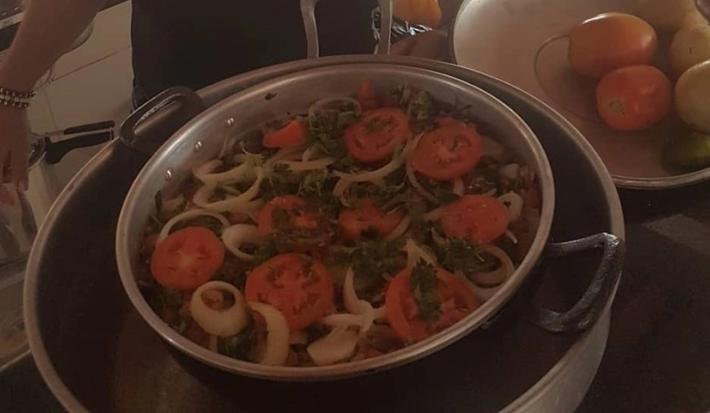 Fotografia de uma panela grande com moqueca com tomates, pimentões e cebolas em rodelas por cima. A panela está dentro de uma outra panela maior.