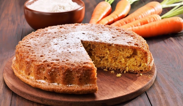 Bolo de cenoura recém-assado em uma tábua de madeira. Com uma crosta dourada e polvilhado com açúcar de confeiteiro, o bolo está parcialmente fatiado, cercado por ingredientes como farinha e cenouras.