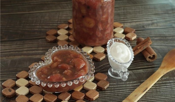 Fotografia em tons escuros de uma mesa de madeira escura. Sobre a mesa tem dois apoios quadriculados de madeira, apoiado em um deles tem um pote de vidro com doce de banana. No outro apoio tem o doce num pote de vidro pequeno em formato de coração.