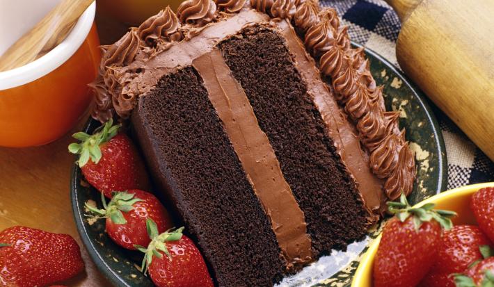 Fotografia de uma fatia de bolo com massa e recheio de chocolate, morangos por toda a parte em um prato desenhado de cor escura, apoiado em uma toalha de mesa azul e branca, quadriculada. Ao lado do bolo, dois potes e um rolo de massa.