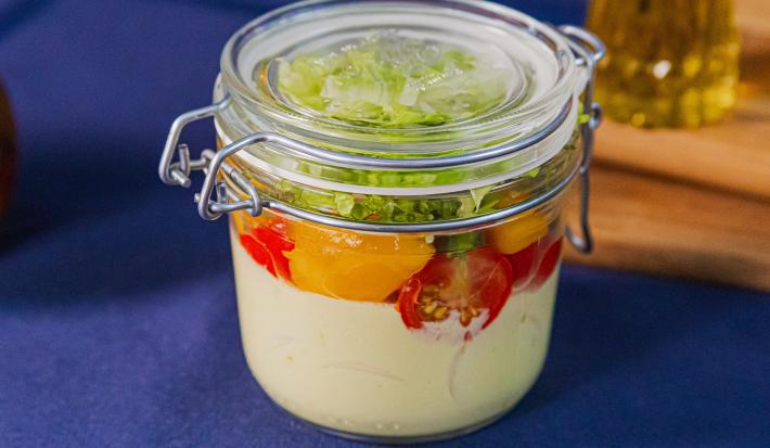 Fotografia em tons de azul com um pote de vidro ao centro. Dentro do pote existe uma salada feita com manga, alface, tomate cereja e molho de mostarda com iogurte