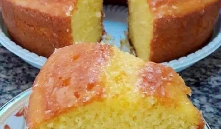 Fotografia de um pedaço grande de bolo de fubá sobre um prato pequeno branco, um pouco fundo, com desenhos delicados. No fundo tem o restante do bolo, quase inteiro, sobre um apoio de plástico branco.