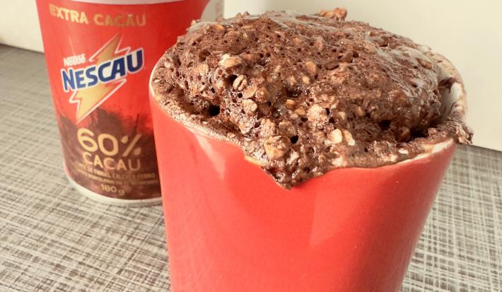 Fotografia de uma caneca vermelha com bolo de chocolate com grãos de aveia. Ao fundo tem uma embalagem de nescau 60% cacau.